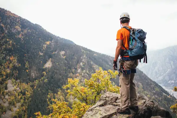 A climber in a helmet and harness, with a pack on, stands on a rock outcrop above a forested valley