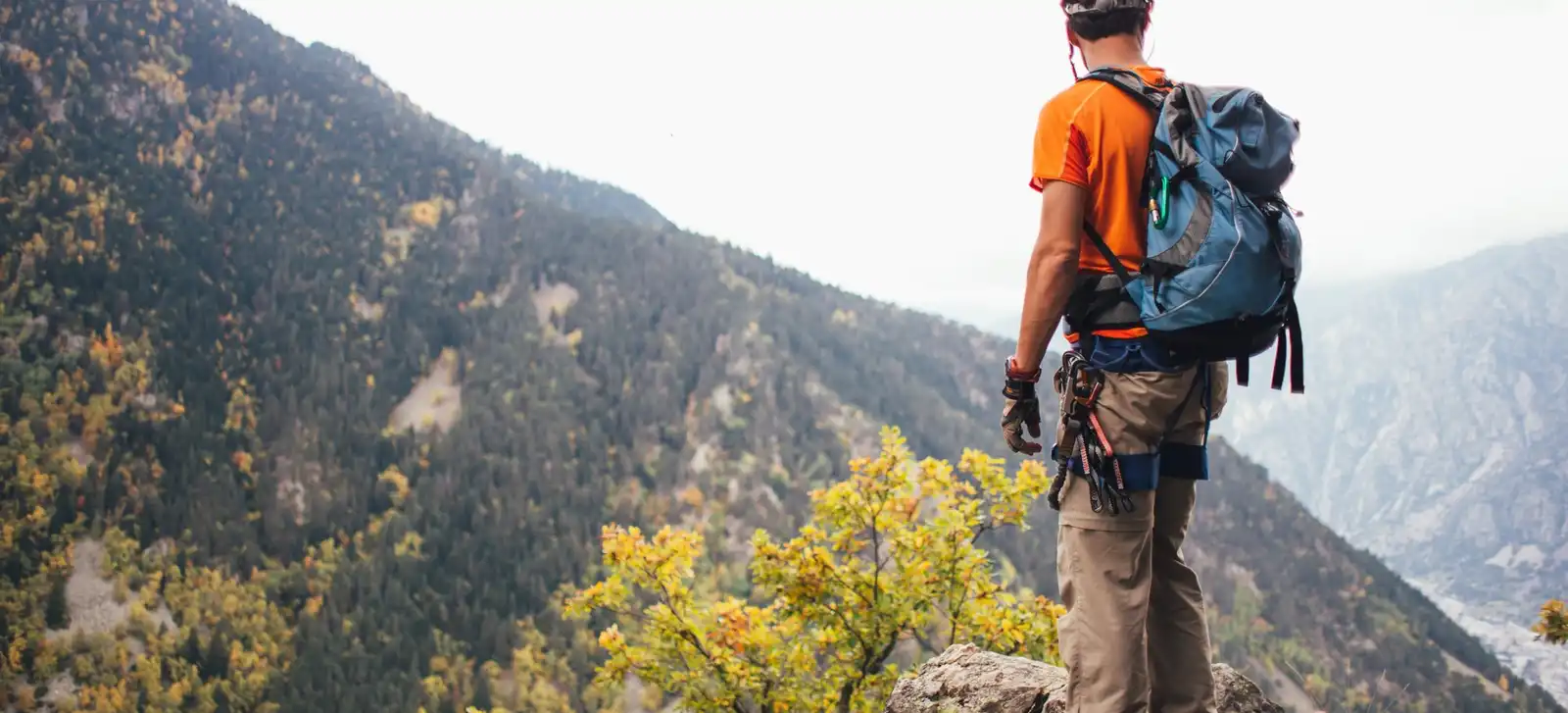 A climber in a helmet and harness, with a pack on, stands on a rock outcrop above a forested valley