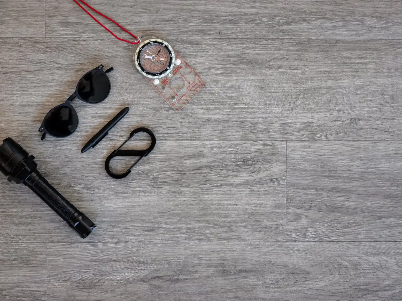 A baseplate compass on a red cord, sunglasses, a pen, a double clip and a torch laid out on grey boards