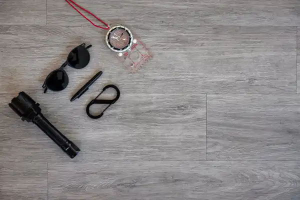 A baseplate compass on a red cord, sunglasses, a pen, a double clip and a torch laid out on grey boards
