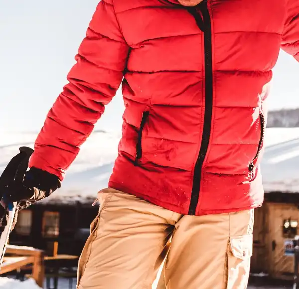 A skier in a red quilted insulated jacket with a black zip and khaki trousers, holding a ski pole