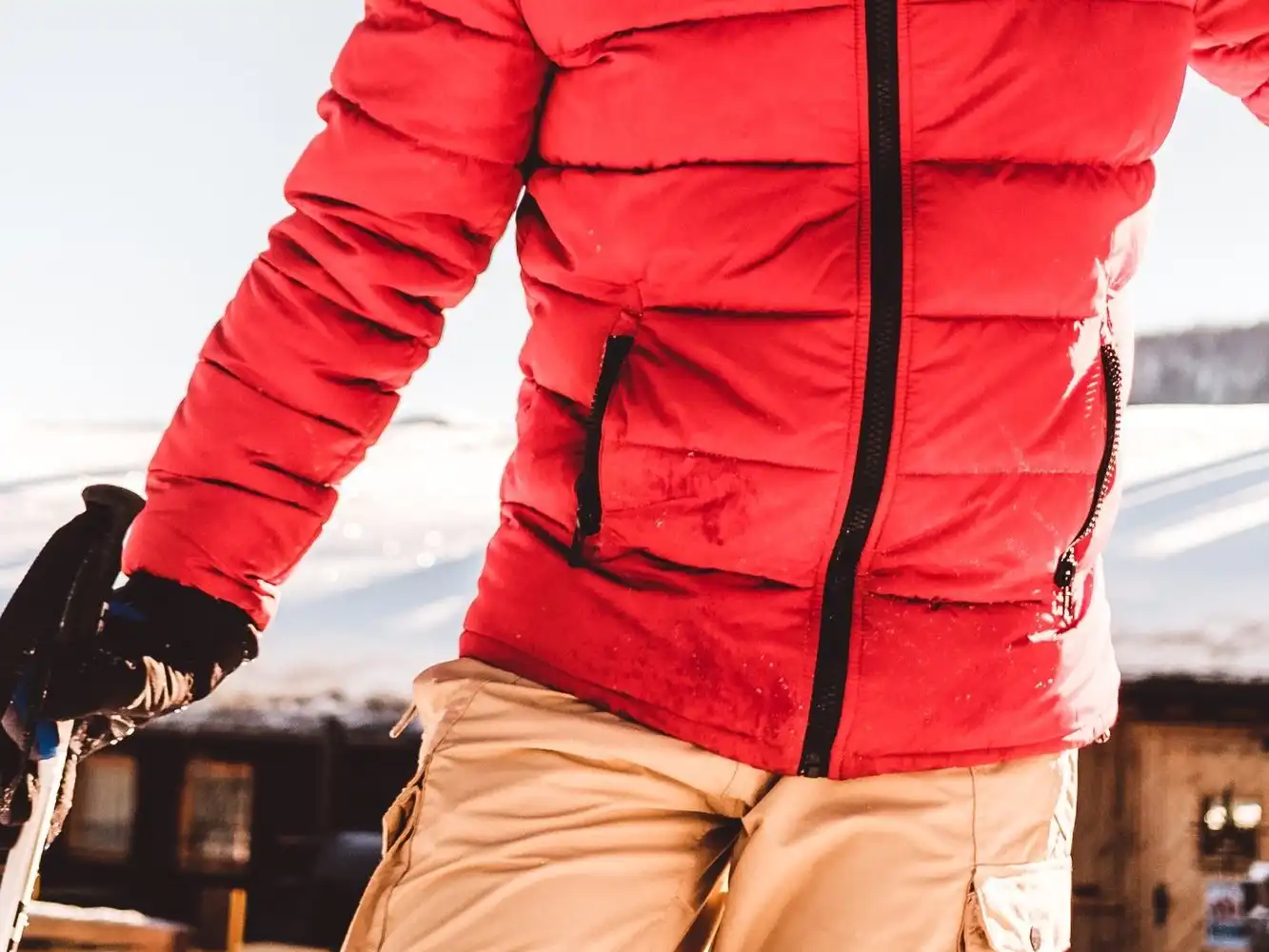 A skier in a red quilted insulated jacket with a black zip and khaki trousers, holding a ski pole