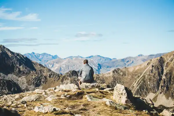A hiker sits on a rocky ridge looking out over a wide valley and rows of mountains under a clear sky