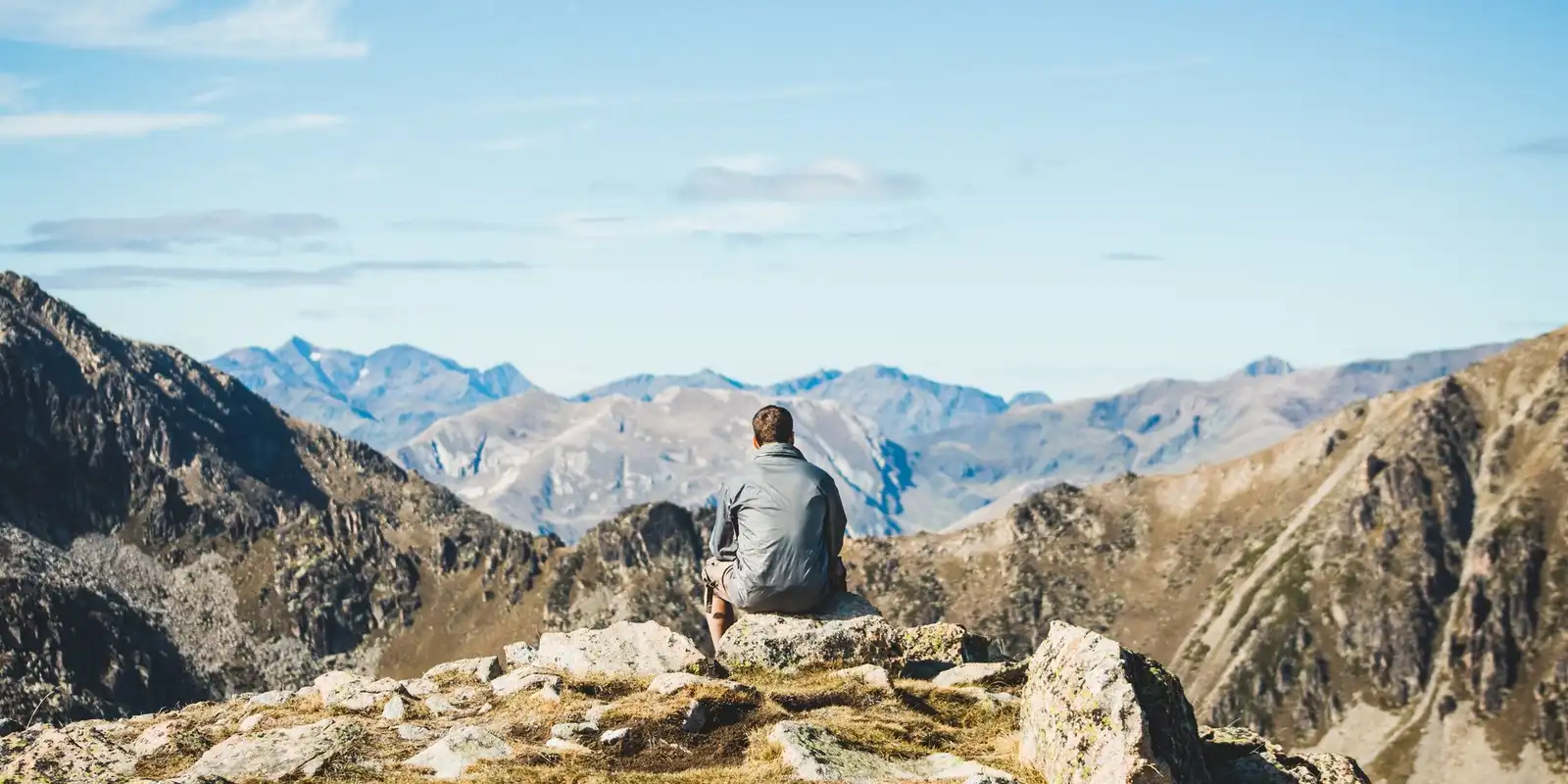 A hiker sits on a rocky ridge looking out over a wide valley and rows of mountains under a clear sky