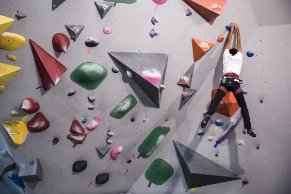 A climber reaches for holds on an indoor bouldering wall covered in coloured volumes
