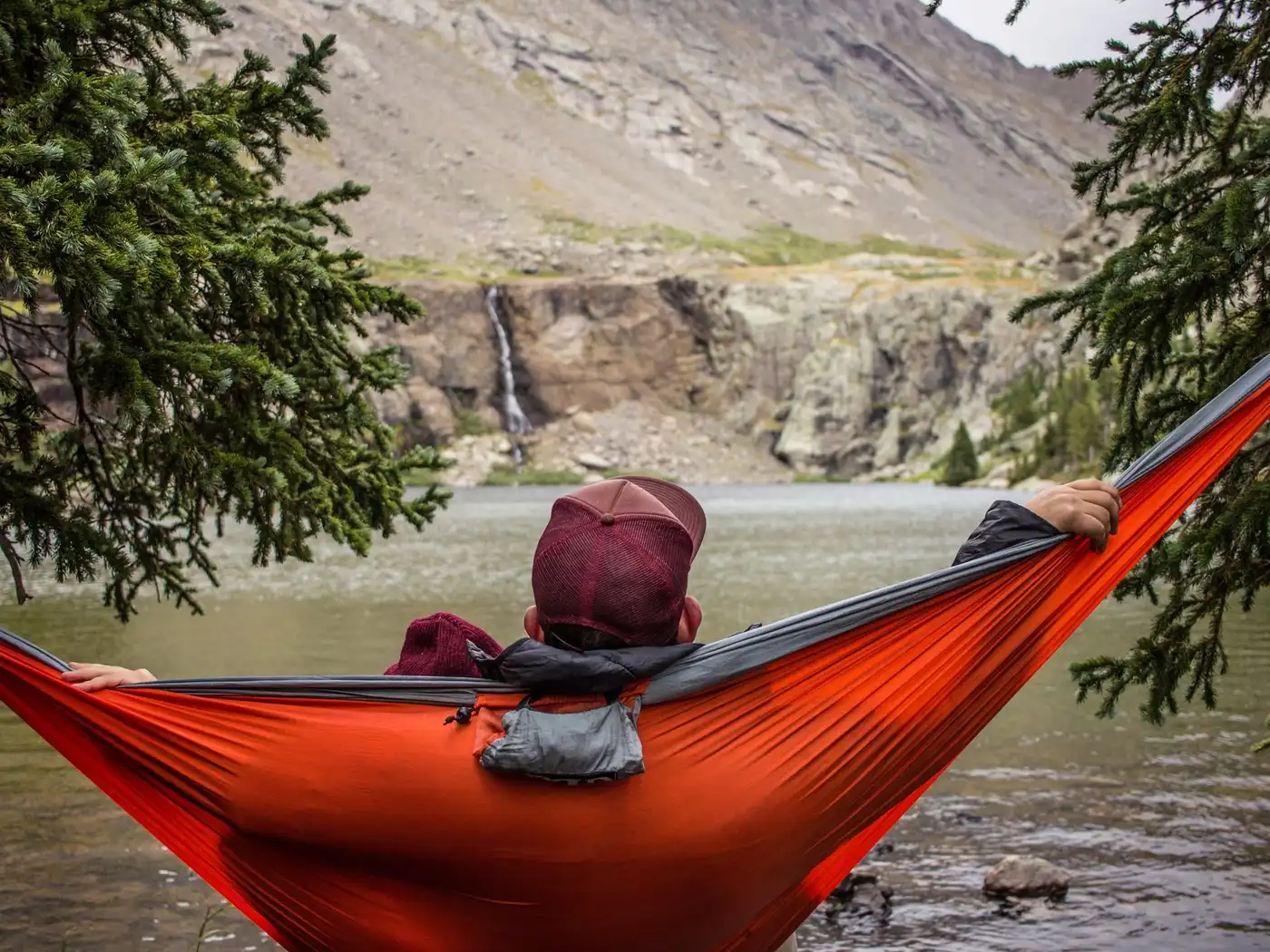 A person lies in an orange hammock strung between pines at the edge of a mountain lake