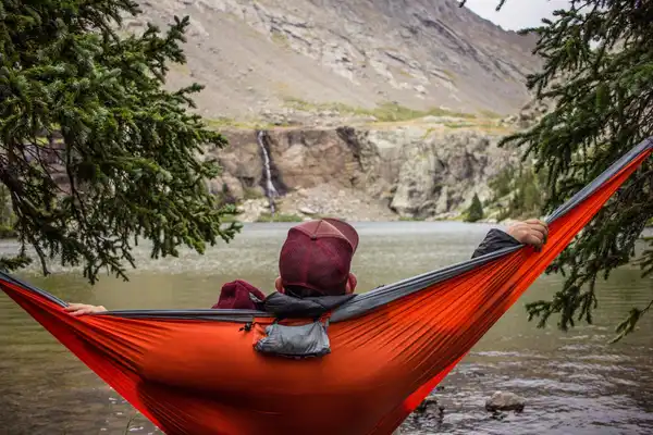 A person lies in an orange hammock strung between pines at the edge of a mountain lake