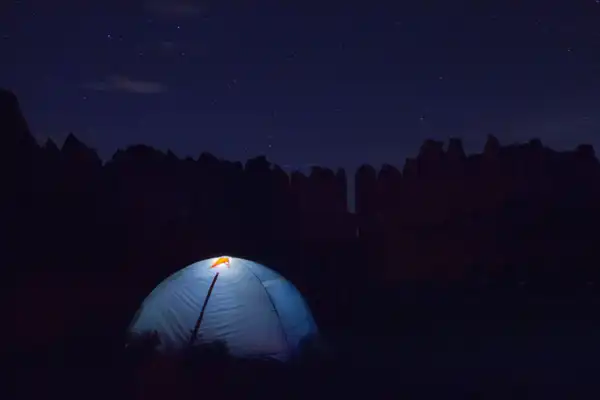 A small blue dome tent glowing from a lamp inside, pitched below dark cliffs under a night sky