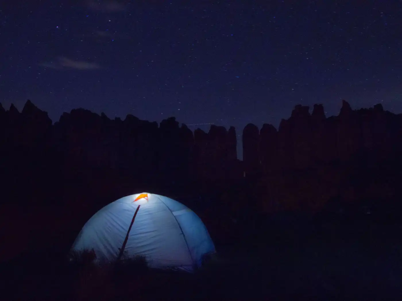 A small blue dome tent glowing from a lamp inside, pitched below dark cliffs under a night sky
