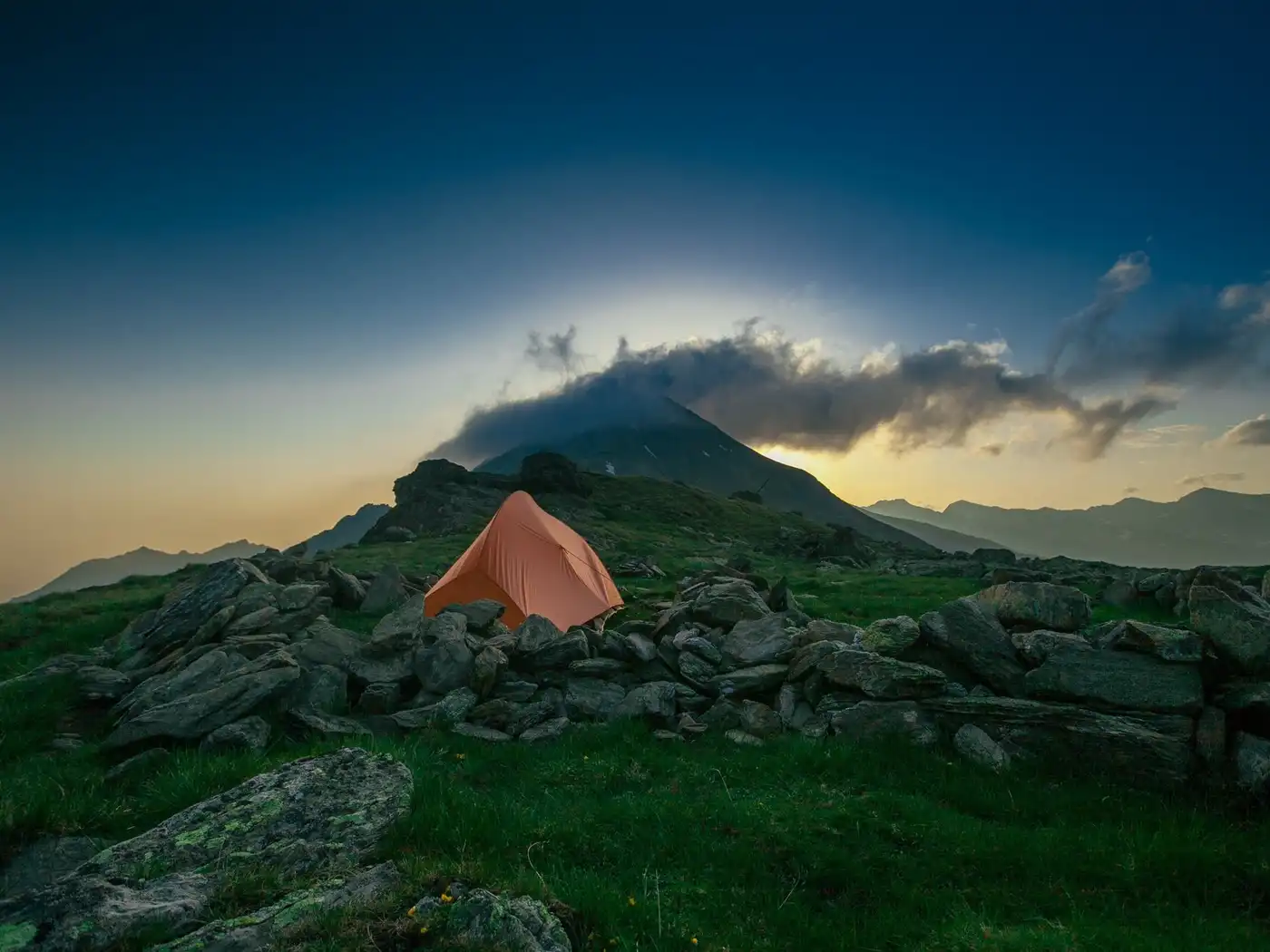 An orange two-person tent pitched among boulders on a green hillside below a cloud-capped peak at dusk