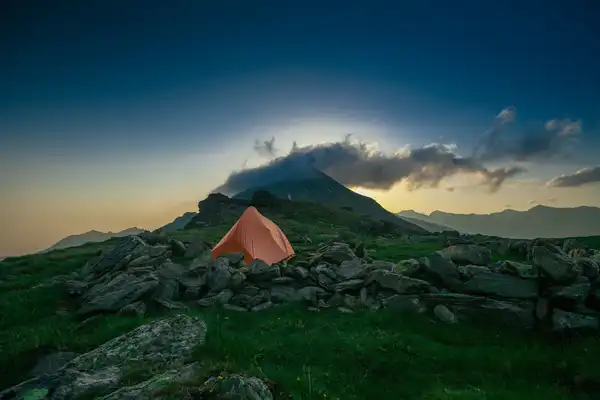 An orange two-person tent pitched among boulders on a green hillside below a cloud-capped peak at dusk