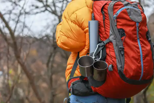 A hiker in a yellow down jacket seen from behind, wearing a red and grey backpack with two steel mugs clipped to the side